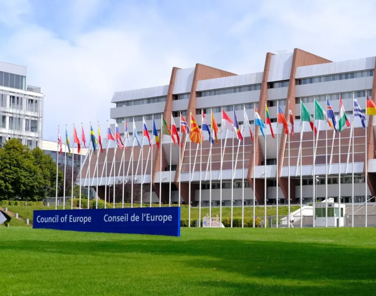 Edificio del Consiglio d'Europa con numerose bandiere esposte davanti, su un'area verde ben curata e il cartello "Council of Europe" visibile in primo piano