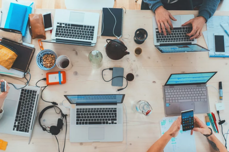 Tavolo di lavoro con laptop, smartphone, cuffie e altri oggetti, dove diverse persone stanno lavorando e utilizzando dispositivi elettronici