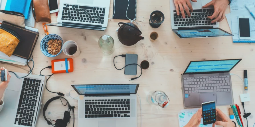 Tavolo di lavoro con laptop, smartphone, cuffie e altri oggetti, dove diverse persone stanno lavorando e utilizzando dispositivi elettronici