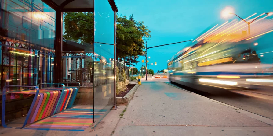 Fermata dell'autobus con panchina colorata e strisce sul pavimento, mentre un autobus in movimento crea un effetto di sfocatura. Il cielo al tramonto aggiunge una luce morbida all'immagine