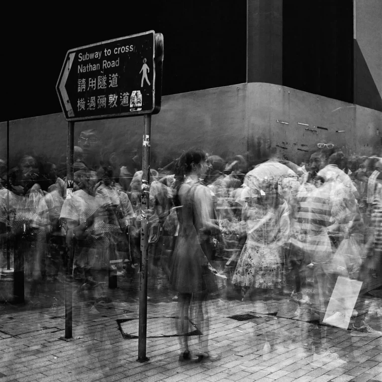 Immagine in bianco e nero di una strada affollata con persone in movimento sfocate, con un cartello che indica un sottopassaggio per attraversare Nathan Road